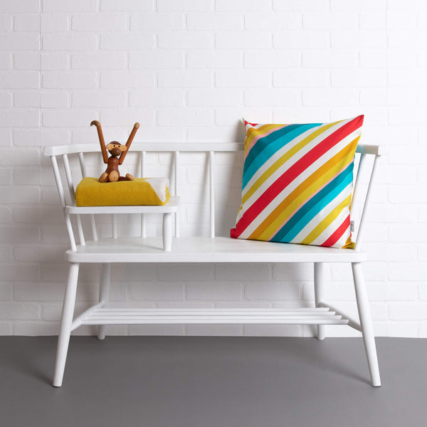 tullibee milo retro diagonal varying width stripe cushion in tones of aqua, red, pink, mustards & white colours on a white ercol telephone bench along with a tullibee picalilli knitted blanket & wooden monkey.   In front of a white brick wall