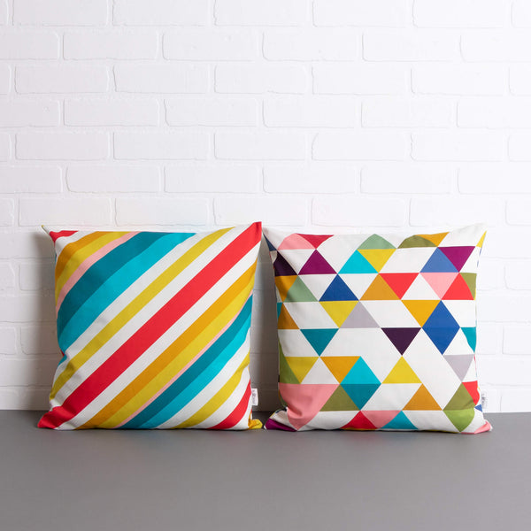 tullibee milo retro diagonal varying width stripe cushion & Yoshi triangle geometric cushion sat side by side on a concrete floor in front of a white brick wall