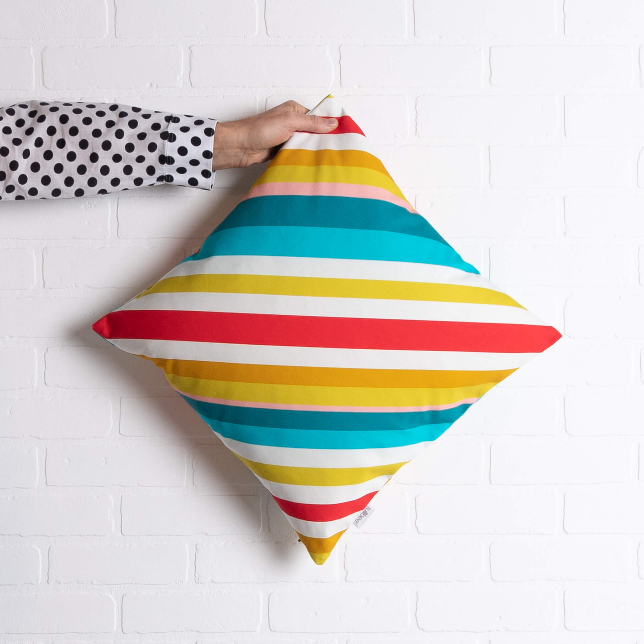 tullibee milo retro diagonal varying width stripe cushion in tones of aqua, red, pink, mustards & white colours being held out by an arm with a black & white spotty shirt