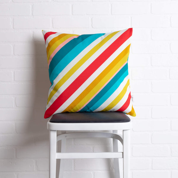 tullibee milo retro diagonal varying width stripe cushion in tones of aqua, red, pink, mustards & white colours sat on a stool in front of a white brick wall