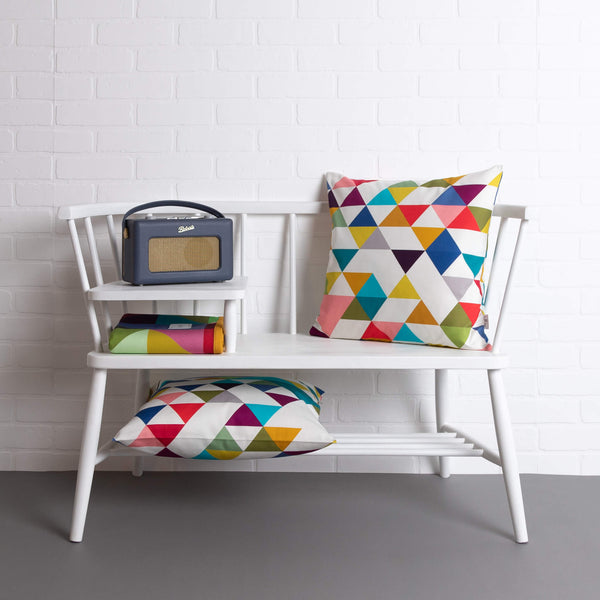 tullibee Yoshi rainbow triangle geometric print cushion on a white ercol telephone bench along with a second Yoshi cushion under bench, colourful tullibee blanket & Roberts retro style radio.   In front of a white brick wall